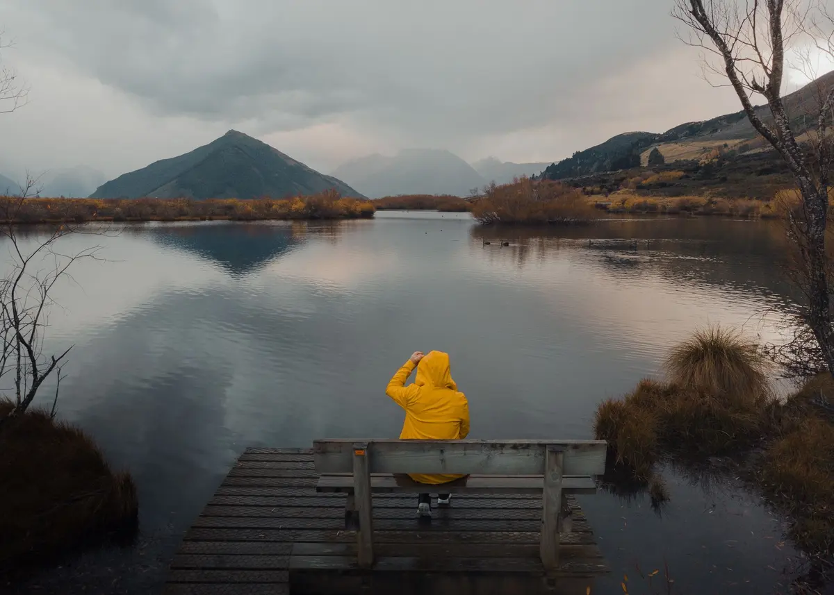 Traveller in a yellow raincoat on a lakeside jetty at Glenorchy, New Zealand, looking out over the lake and mountains
